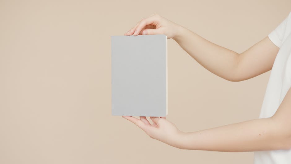 Hands holding a blank book cover for creative design mockup on a neutral background.