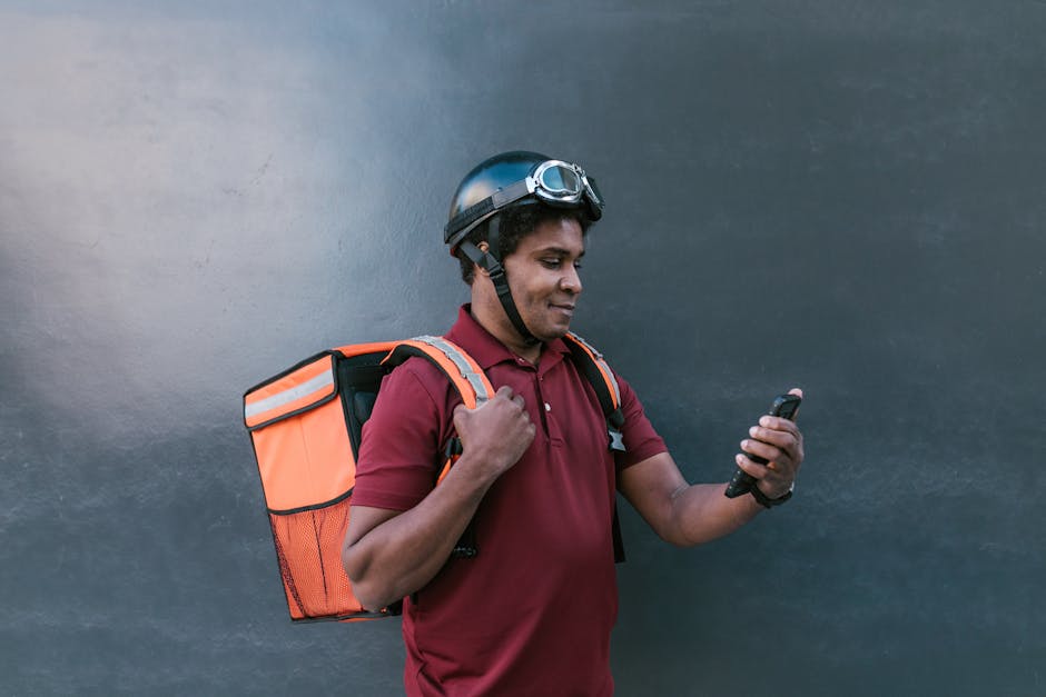 A delivery courier wearing a helmet and backpack checks his smartphone against a dark background.